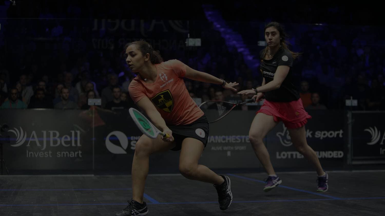 Two professional squash players mid-rally on a glass show court under tournament lighting